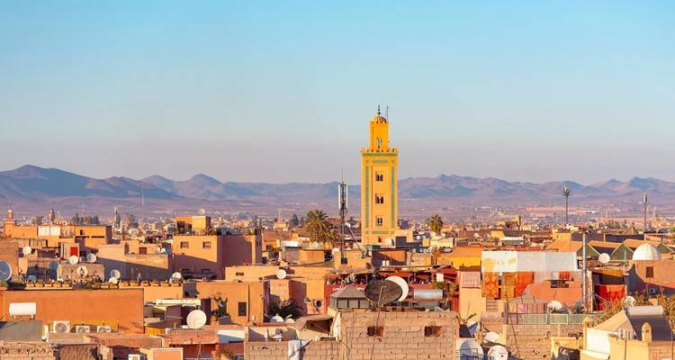 Panoramablick auf die Skyline von Marrakesch mit dem Minarett der Koutoubia-Moschee, das sich über sonnenbeschienene Dächer und entfernte Berge erhebt.