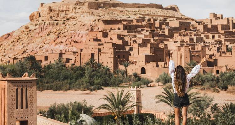 Reisender mit erhobenen Armen genießt den Blick auf das aus Lehm erbaute Ksar von Aït Benhaddou, umgeben von Wüstenpalmen.
