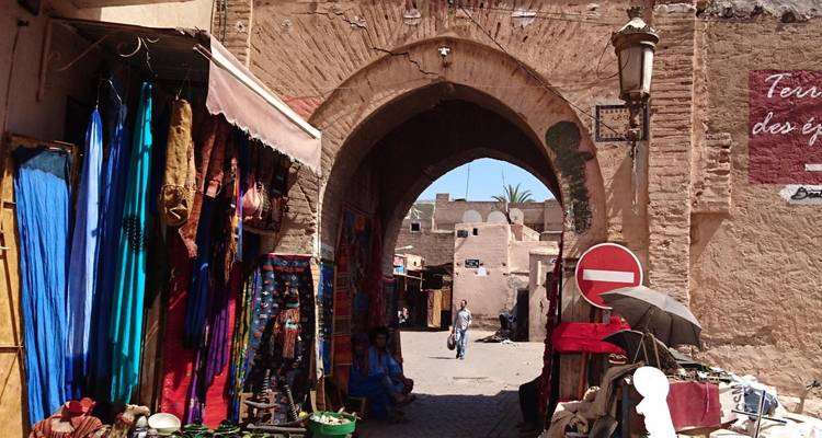 Schmaler Marktplatzgasse unter einem Steinbogen in der Medina von Marrakesch, gesäumt mit Stoffen und Waren.