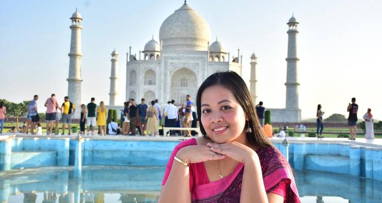Mujer sonriente con un sari rosa posando con las manos bajo la barbilla, el Taj Mahal y un estanque de reflexión detrás.