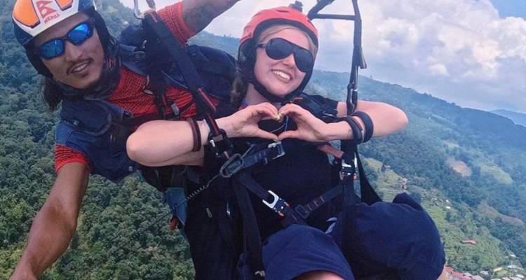 Tandem paragliding pair high above lush green hills, the woman forming a heart with her hands.