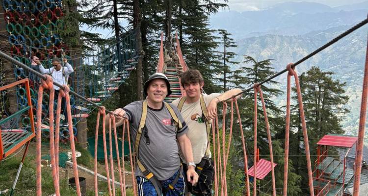 Two men smiling on a high rope bridge adventure course amid tall conifer trees and mountain views.