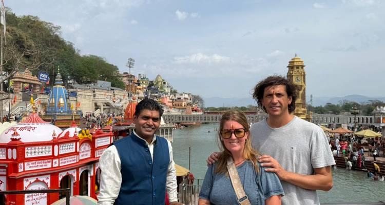 Zwei Reisende und ihr Führer stehen neben dem Ganges am Har Ki Pauri ghat, mit dem ikonischen Uhrturm und Tempeln im Hintergrund.