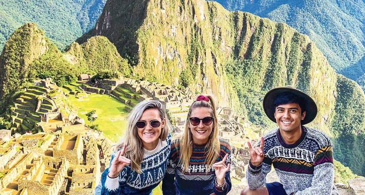 Tres viajeros sonrientes en suéteres estampados posan con las icónicas terrazas de Machu Picchu detrás de ellos.