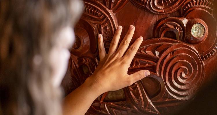 Primer plano de la mano de un visitante tocando un panel de madera roja maorí intrincadamente tallado bajo la suave iluminación del museo.