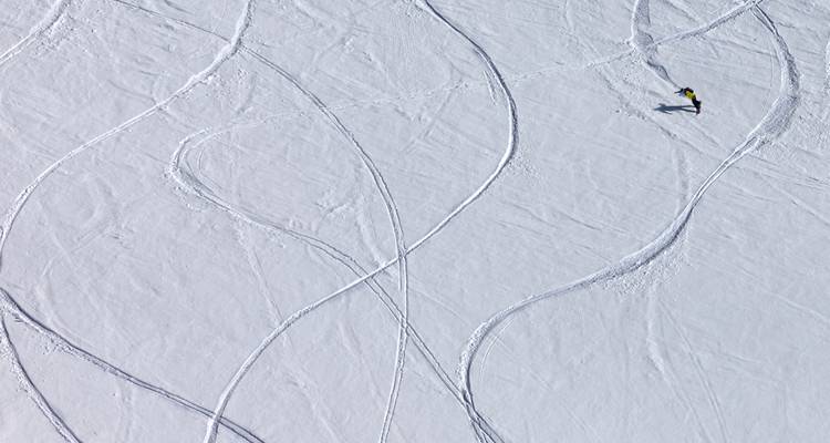 Bovenaanzicht van verse skisporen die patronen in ongerepte sneeuw uithakken met een eenzame skiër in felle kleding.