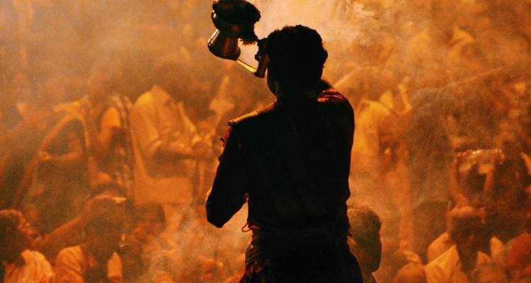 Un sacerdote recortado contra la luz realiza un ritual de incienso entre el humo que se arremolina durante una ceremonia fluvial al atardecer.