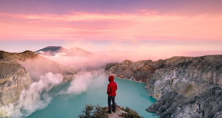 Adventurer in red jacket admiring steamy turquoise lake inside Ijen crater under pink dawn sky.