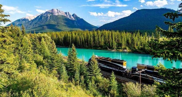 Trein bij een turquoise rivier te midden van een bebost berglandschap.