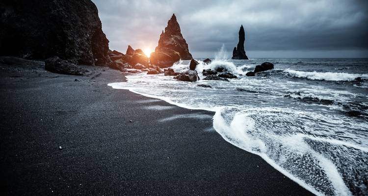 Dramatisch zwart zandstrand met rotsachtige zeerotsen bij zonsondergang.