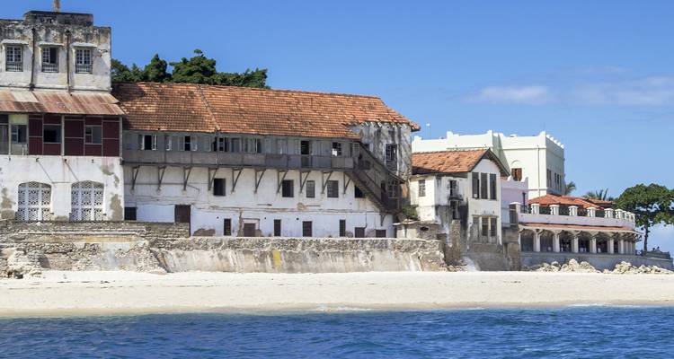 Edificios históricos en el frente de agua con fachadas desgastadas alineados a lo largo de una playa arenosa y el mar azul en Stone Town.