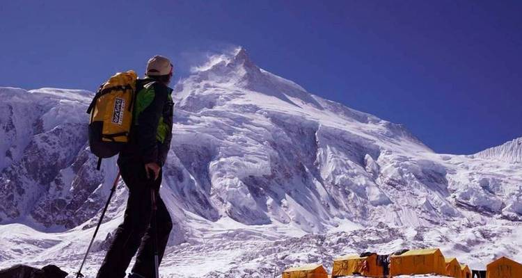 Person in outdoor gear looking at a snowy mountain peak with tents below.