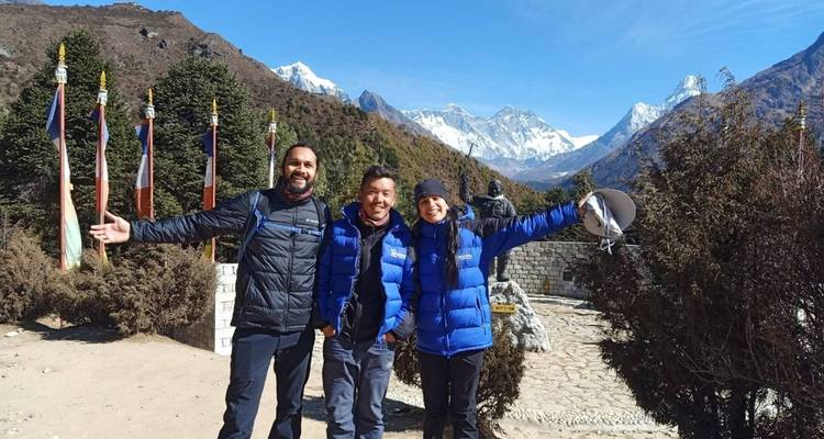Drei Personen mit ausgestreckten Armen, Berge und Flaggen im Hintergrund sichtbar.
