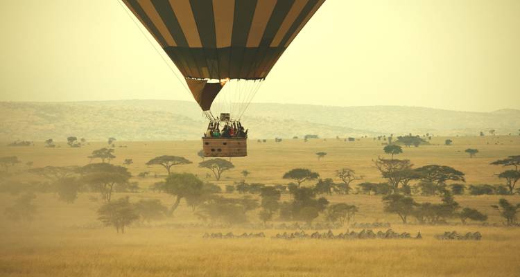 Heißluftballon mit Passagieren über einer weiten Savannenlandschaft.