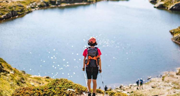 Excursionista con bastones de senderismo contempla un tranquilo lago alpino rodeado de laderas rocosas.