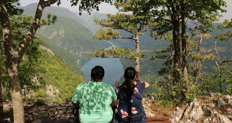 Dos personas sentadas en un banco con vista a un valle pintoresco y un río.