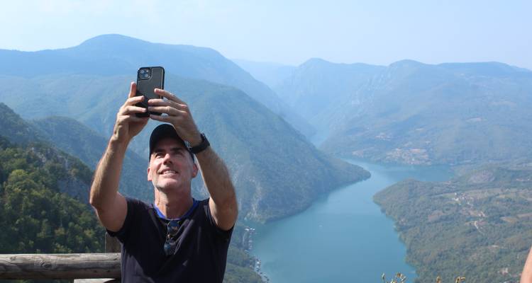 Hombre tomándose una selfie con un valle pintoresco y un río de fondo.