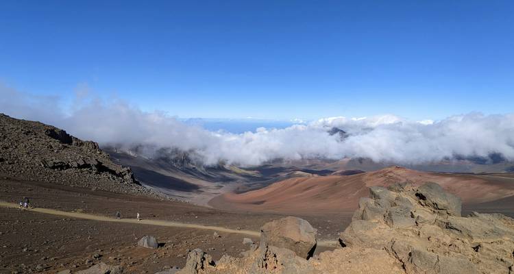 Vue panoramique sur un paysage de cratère volcanique avec des crêtes rocheuses, une terre rouge-brun et une mer de nuages sous un ciel bleu dégagé à Hawaii.