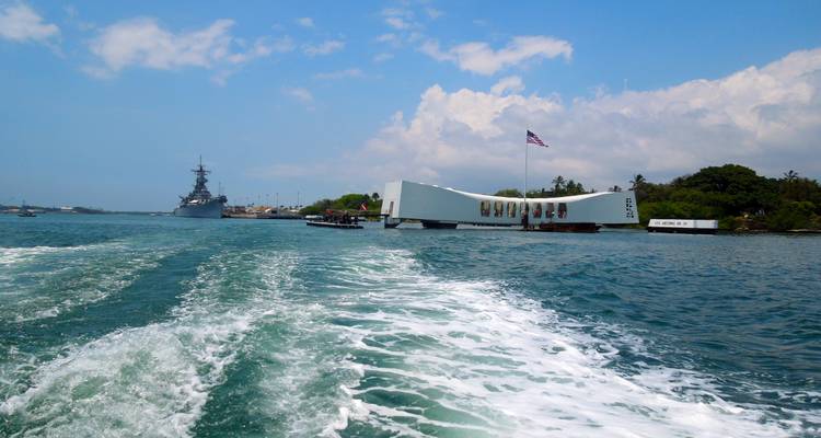 Vue depuis un bateau du mémorial de l'USS Arizona à Pearl Harbor avec un drapeau américain flottant au-dessus d'une eau bleue calme.