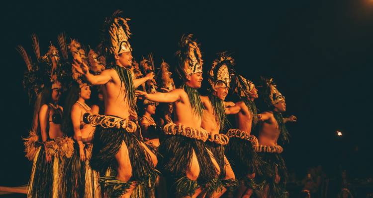 Des danseurs polynésiens traditionnels vêtus de jupes en herbe et de coiffures ornées qui se produisent avec énergie sur une scène sombre.