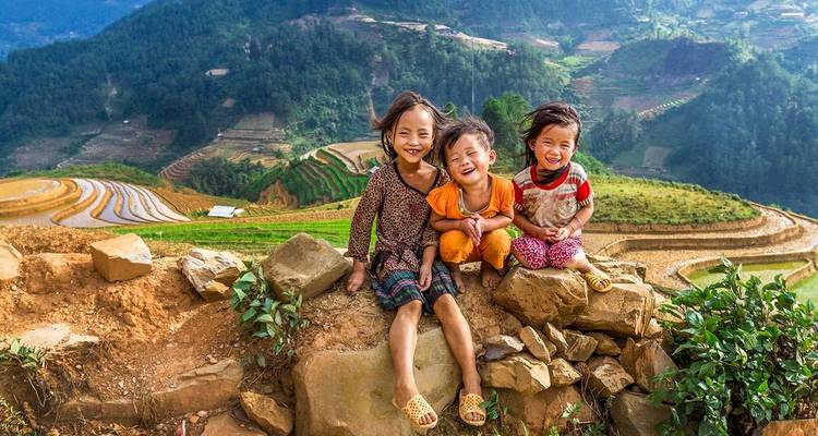 Three laughing children sit on a stone wall overlooking emerald rice terraces in Sapa.