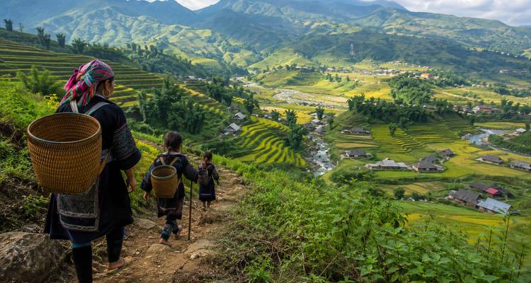 A woman and two children in traditional dress hike down a dirt path overlooking terraced rice fields and a mountain valley.