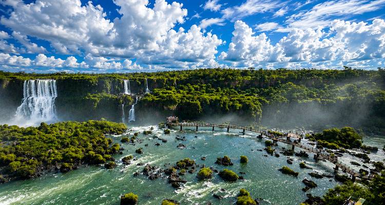 Vista aérea panorámica de las Cataratas del Iguazú con selva tropical exuberante, puente pasarela sobre río turbulento, y cielo dramático lleno de nubes.