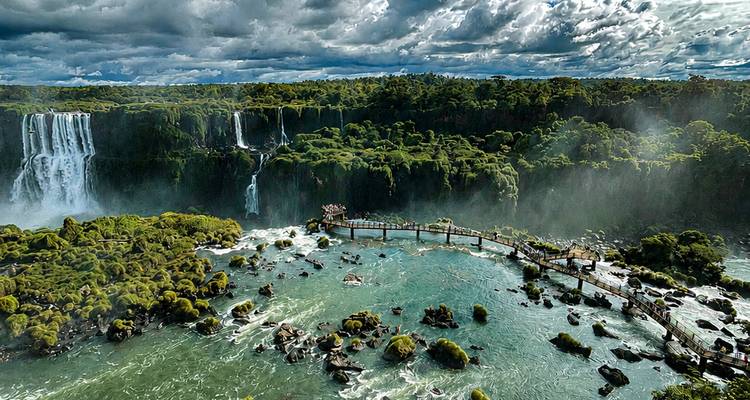 Vista amplia de las Cataratas del Iguazú enmarcadas por nubes tormentosas, bosque esmeralda y niebla que se eleva de las aguas agitadas.