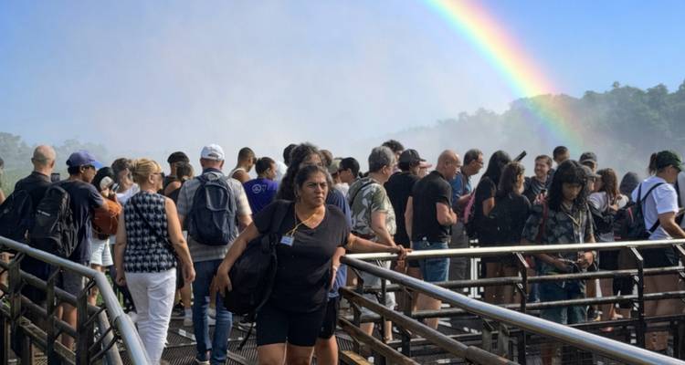 Gran multitud en una pasarela de metal en las Cataratas del Iguazú con un arco iris vibrante formándose en la bruma de spray.