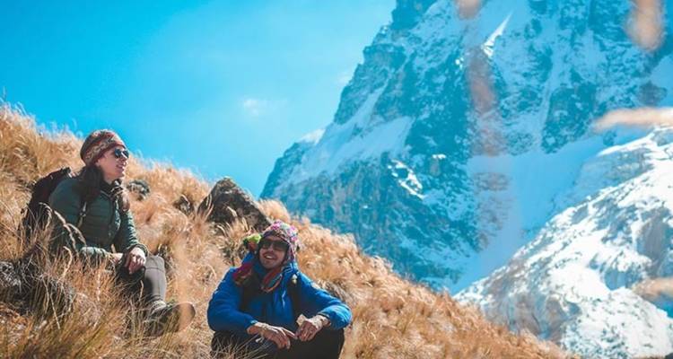 Dos excursionistas descansando en una ladera cubierta de hierba con picos nevados en el fondo.