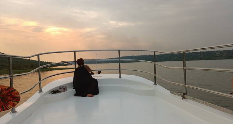Un viajero con capa sentado en la proa de un barco blanco navegando por un río tranquilo al atardecer.