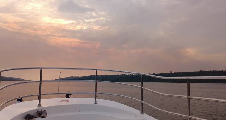 Vista desde la proa de un bote navegando en un lago o río ancho bajo un cielo vespertino brumoso con colinas verdes a lo lejos.