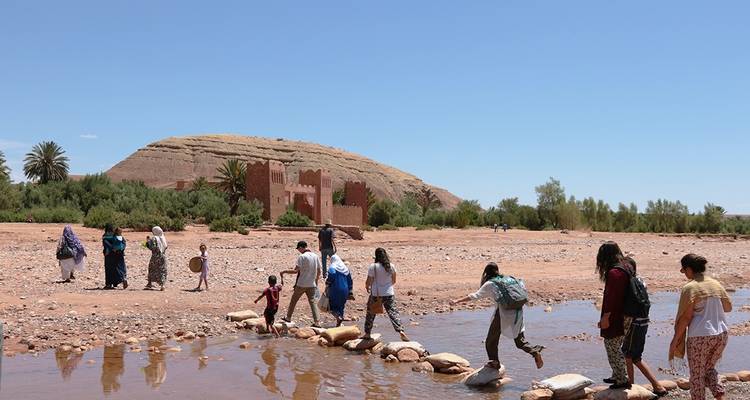 Menschen überqueren einen seichten Fluss mit einem historischen Gebäude im Hintergrund.