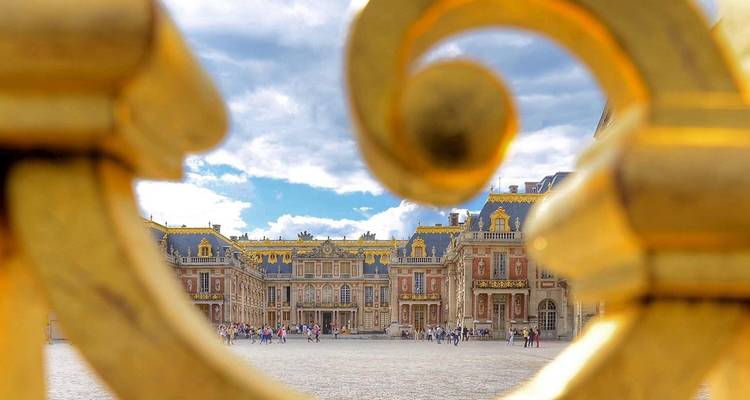 Vue des cours du Château de Versailles cadrée de manière créative à travers un portail doré ornementé.