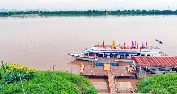 A passenger boat with colorful flags is docked at a quiet riverside pier surrounded by greenery.