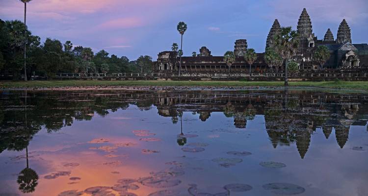 Angkor Wat and its reflection glow at dawn beside a still lotus pond under pastel skies.