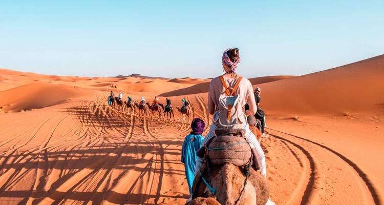 Kamelenkaravaan met reizigers die over golvende oranje duinen trekken onder een strakblauwe hemel.