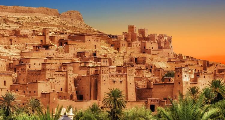 Zonsondergang boven de aarden kasbahs van Ait Benhaddou, omgeven door palmbomen en een levendige hemel.