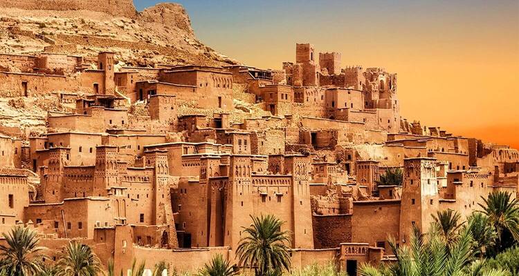 Goudkleurige zonsondergang boven de klei-vestingstad Ait Benhaddou met palmbomen op de voorgrond.