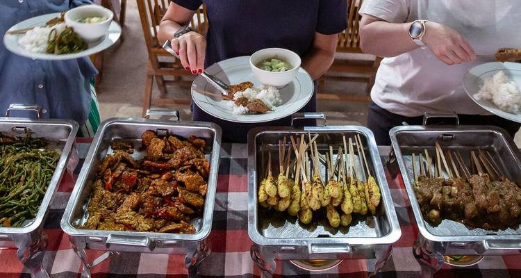 Buffet de plats indonésiens comprenant des brochettes satay et du tempeh disposés dans des chauffe-plats en métal.