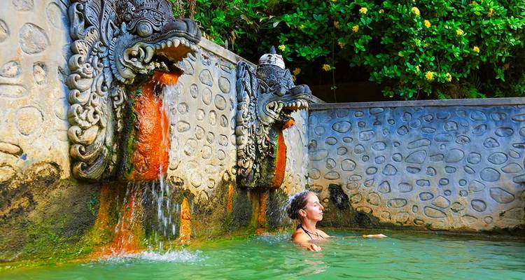 Un voyageur se détend dans un bassin de source chaude alimenté par d'ornementales fontaines de dragons en pierre avec une végétation luxuriante à proximité.