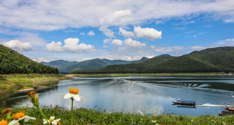 Un lago tranquilo rodeado de colinas verdes y exuberantes y montañas bajo un cielo azul brillante salpicado de nubes blancas, con un bote de cola larga dejando estelas detrás.