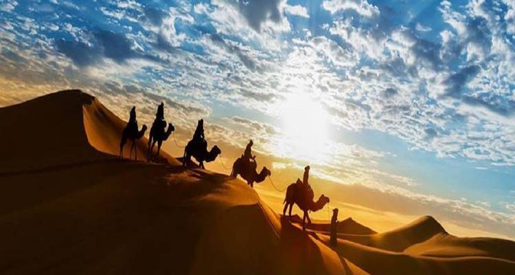 Silhouetten van ruiters op kamelen die een zandduinkam oversteken tegen een dramatische zonsopganghemel