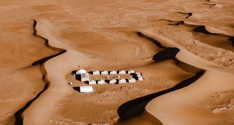 Vista aérea de un campamento remoto en el desierto formado por tiendas blancas ordenadas entre dunas de arena ondulantes.