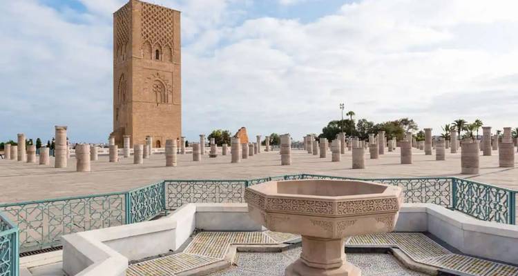 Large plaza with Hassan Tower and surrounding stone columns beside an ornate fountain under soft clouds.