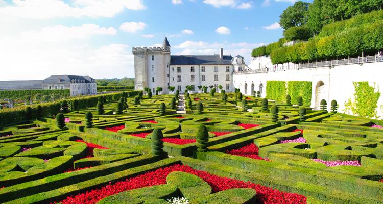 Un parterre géométrique vibrant de haies rouges et vertes s'étend sous un château blanc lors d'un après-midi ensoleillé.