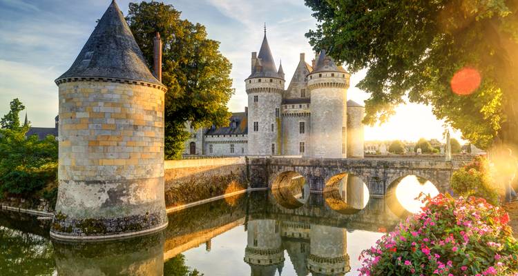 La lumière dorée du soleil illumine un château de pierre entouré de douves avec des tours rondes se reflétant dans une eau tranquille bordée de fleurs.