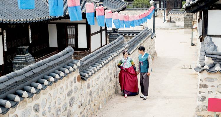 Pareja joven en hanbok colorido caminando junto a los muros de piedra de un callejón tradicional de un pueblo coreano