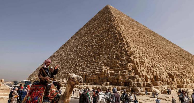 Man op een kameel voor een piramide met talrijke toeristen rondom.