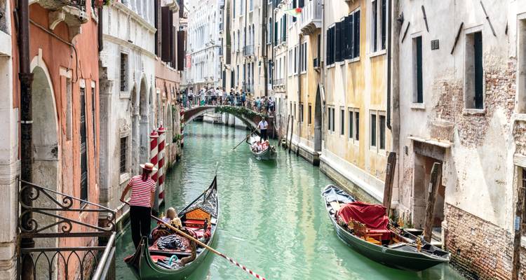Picturesque narrow canal in Venice with gondolas gliding beneath a small pedestrian bridge flanked by colorful facades.